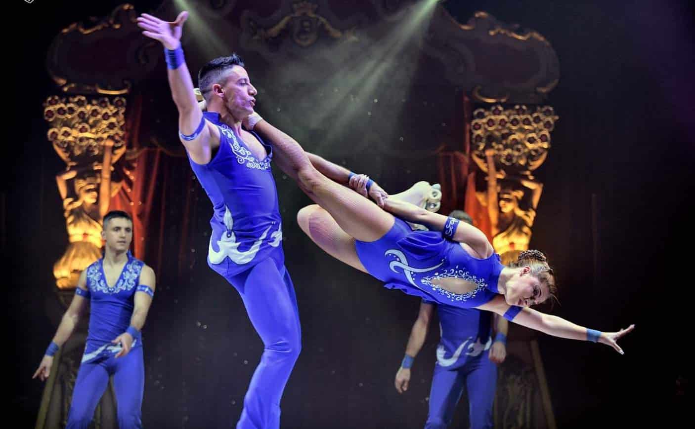 Zwei Artisten führen eine spektakuläre Rollschuh-Akrobatiknummer auf, bei der eine Frau im blauen Kostüm in einer dynamischen Pose in der Luft schwebt, während sie von ihrem Partner, ebenfalls in Blau gekleidet, gehalten wird. Im Hintergrund ist eine festlich beleuchtete Zirkusbühne mit dekorativen Elementen zu sehen.