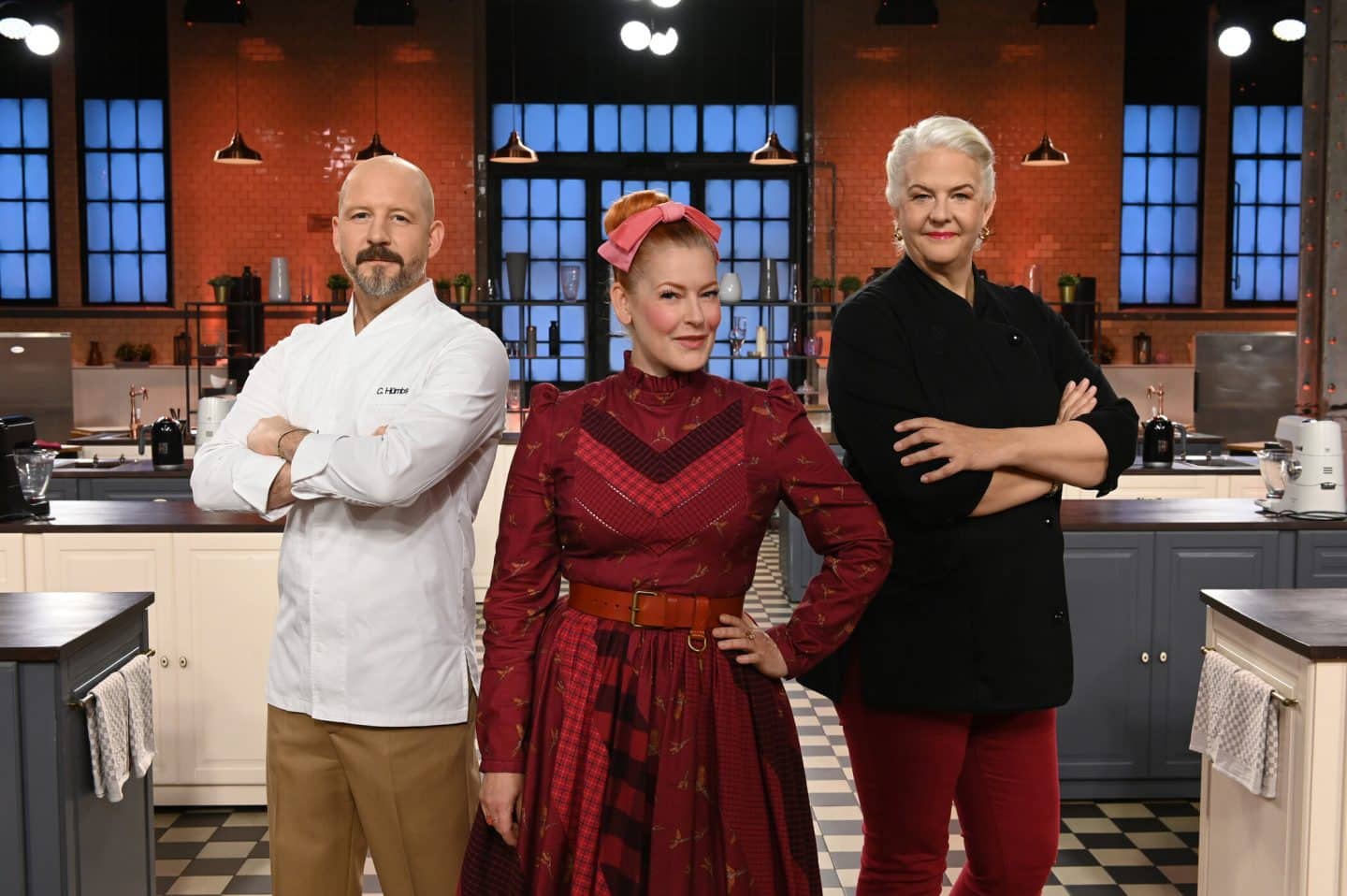 Die Jury der Show mit Christian Hümbs (links), Enie van de Meiklokjes (Mitte) und Bettina Schliephake-Burchardt (rechts) steht in einem stilvoll eingerichteten Studio mit verschränkten Armen. Im Hintergrund sind Küchenutensilien und Arbeitsbereiche zu sehen.