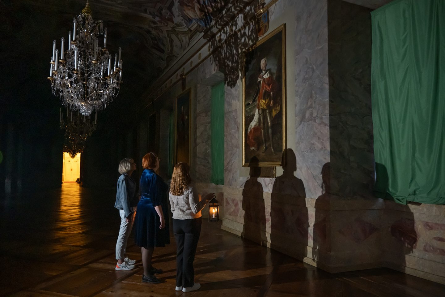 Mehrere Personen betrachten bei gedämpftem Licht ein historisches Gemälde in einem prächtigen Raum des Schlosses Ludwigsburg. Schatten und eine Laterne sorgen für eine geheimnisvolle Atmosphäre.