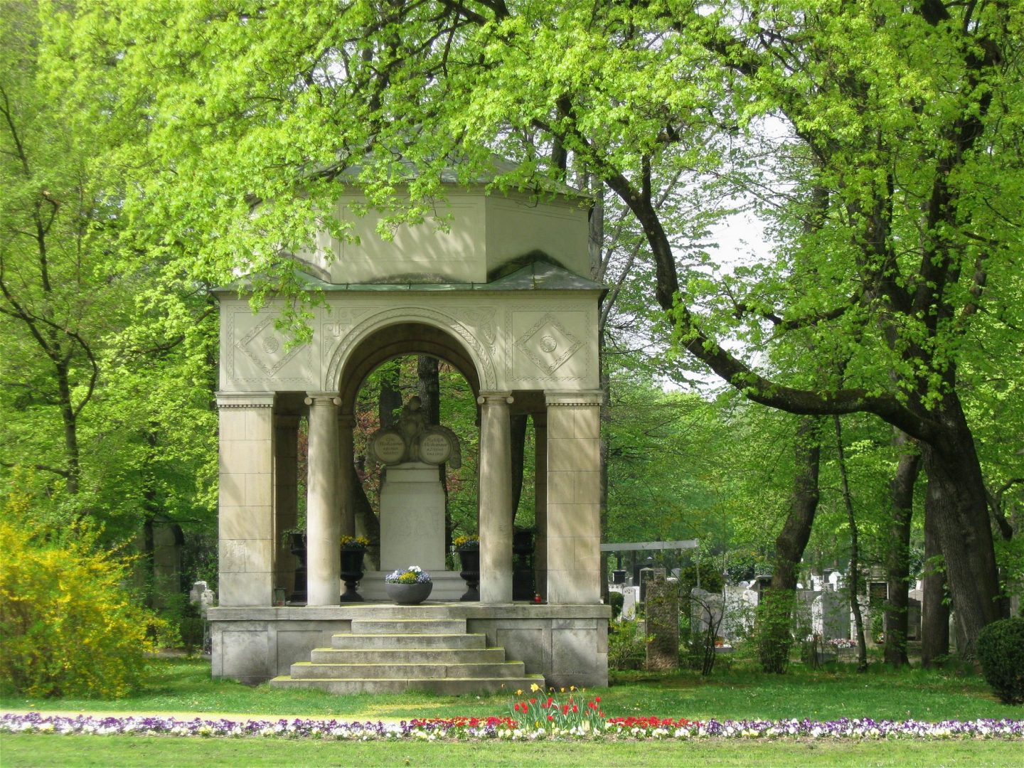 Moshammer-Mausoleum auf dem Münchner Ostfriedhof, umgeben von grünen Bäumen und Blumenbeeten, mit einer eindrucksvollen architektonischen Gestaltung.