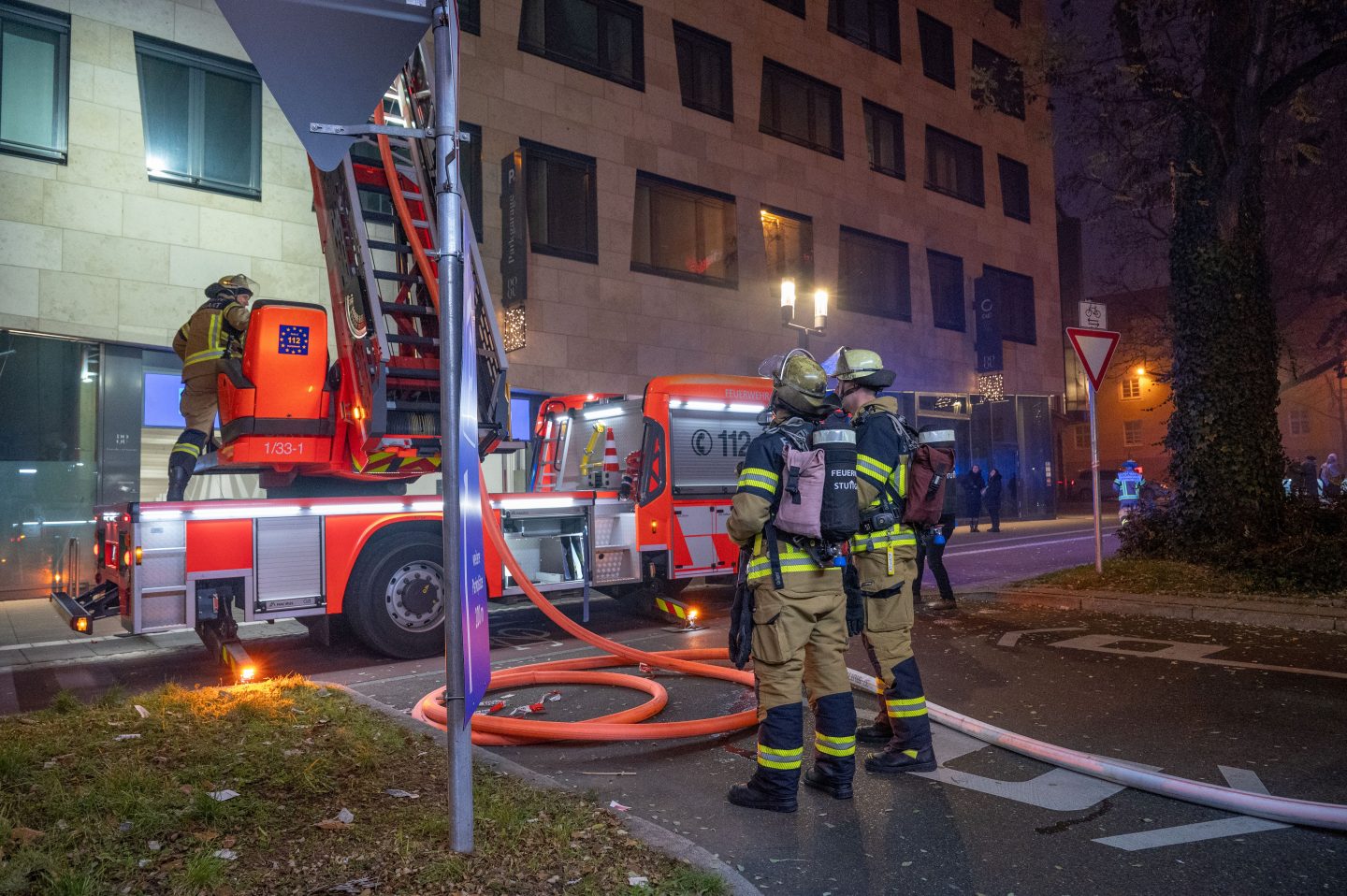 Feuerwehrkräfte in voller Ausrüstung stehen vor einem Einsatzfahrzeug mit ausgefahrener Drehleiter in der Nacht. Im Hintergrund ist ein Gebäude zu sehen, während orangefarbene Schläuche am Boden liegen und die Einsatzkräfte die Szene beobachten.