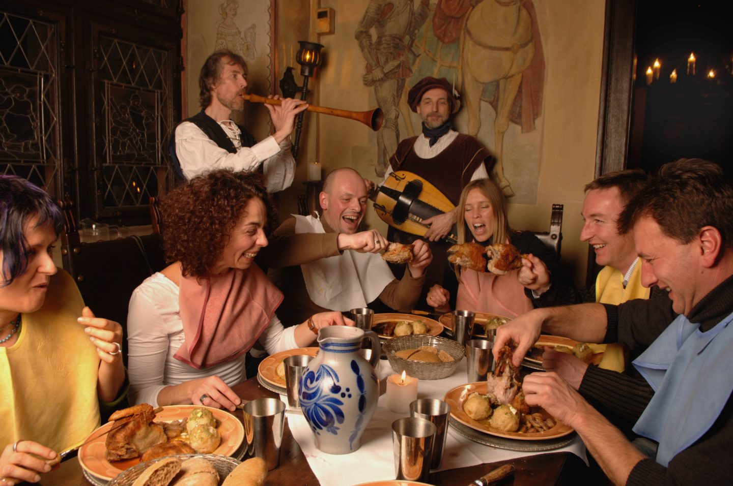 Gäste genießen in mittelalterlicher Kulisse ein festliches "Alemannisches Rittermahl" im Europa-Park. An einer rustikalen Tafel wird herzhaft gespeist, während Musiker in historischen Gewändern mit Flöte und Drehleier für stimmungsvolle Unterhaltung sorgen. Die Atmosphäre wird von Kerzenlicht und Wandmalereien abgerundet.