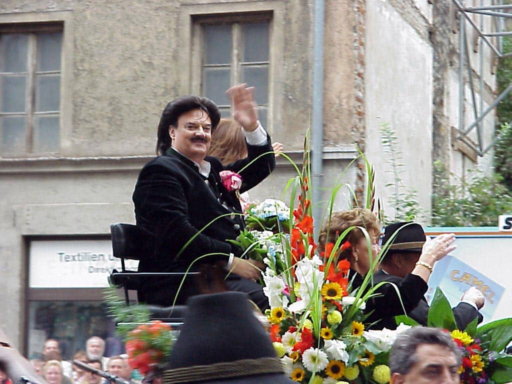 Rudolph Moshammer auf einem geschmückten Festwagen beim Oktoberfest 2000 in München, winkend, umgeben von Blumenarrangements und traditionell gekleideten Personen.