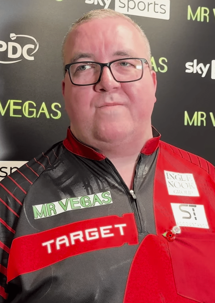 Stephen Bunting, Darts-Profi, in rotem Trikot mit Sponsorenlogos, vor einer Sky-Sports-Wand.