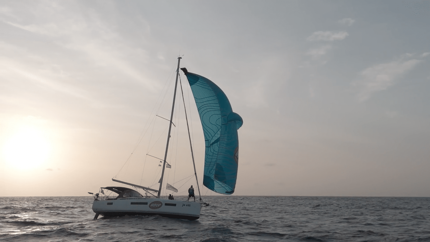 Ein Segelboot aus „MISSION UNKNOWN: Atlantik“ segelt bei Sonnenuntergang über den offenen Ozean. Das Boot trägt ein großes, blaues Segel mit Sponsor-Logos. Die Crew ist an Bord, während sich das Boot durch die Wellen bewegt. Der Himmel ist leicht bewölkt, das Meer zeigt eine sanfte Dünung.

