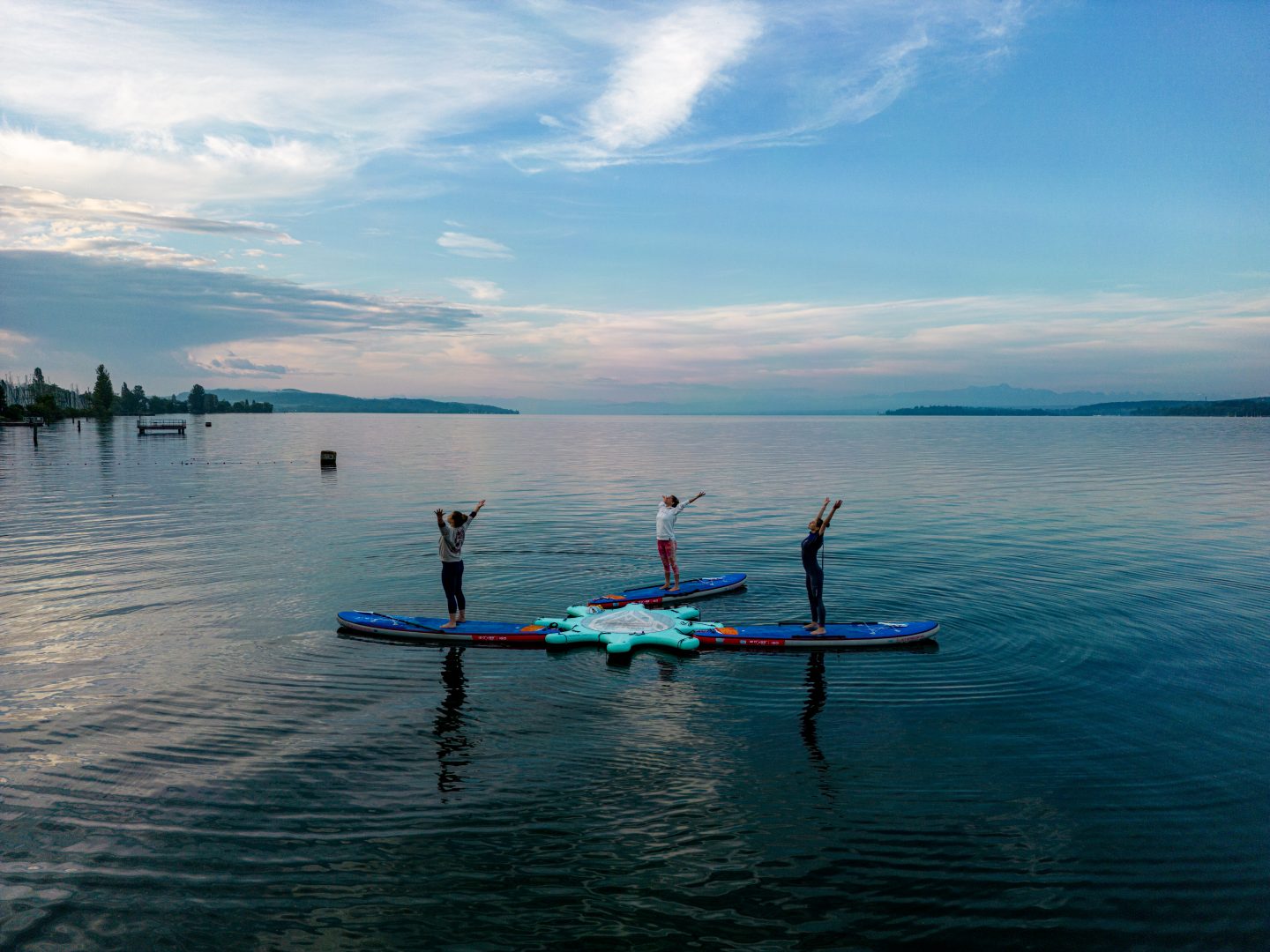 Drei Menschen stehen auf Paddleboards auf einem stillen See und machen Yoga-Übungen. Der Himmel ist leicht bewölkt, und die Uferlinie sowie Berge sind in der Ferne zu sehen.