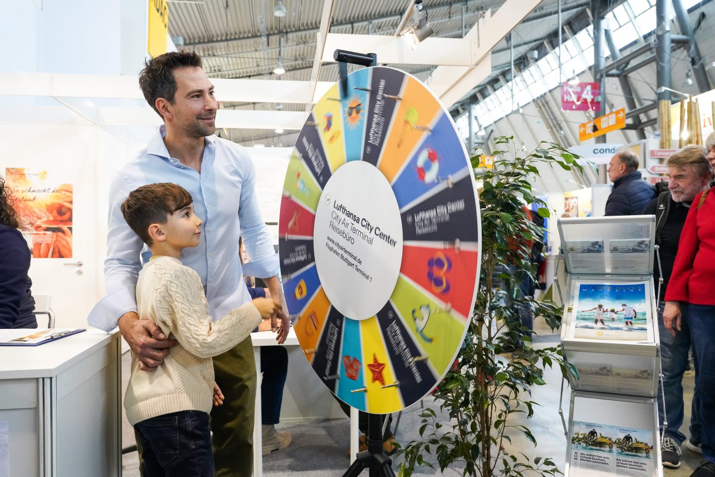 Ein Vater und sein Sohn stehen gemeinsam an einem Glücksrad auf der CMT in Stuttgart. Der Vater hält den Jungen, während dieser das Glücksrad dreht. Im Hintergrund sind weitere Messebesucher und Infostände zu sehen.