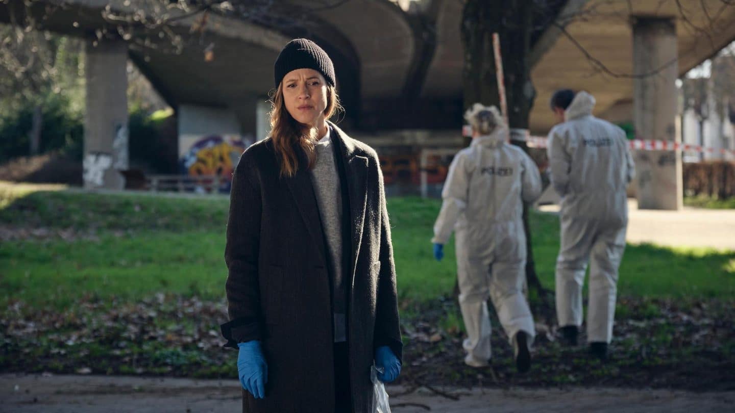 Johanna Stern, die Ermittlerin, steht nachdenklich mit einer schwarzen Mütze und blauen Handschuhen auf einer Wiese unter einer Brücke. Im Hintergrund arbeiten forensische Experten in weißen Schutzanzügen an einem abgesperrten Tatort. Die Atmosphäre ist ernst und angespannt.






