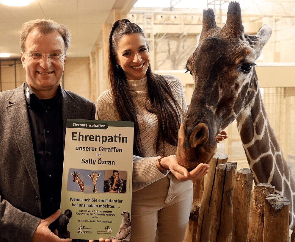 Zoodirektor Dr. Matthias Reinschmidt und Sally Özcan stehen vor einer Netzgiraffe im Zoo Karlsruhe und halten ein Schild, das die Ehrenpatenschaft von Sally Özcan für die Giraffen bekannt gibt.