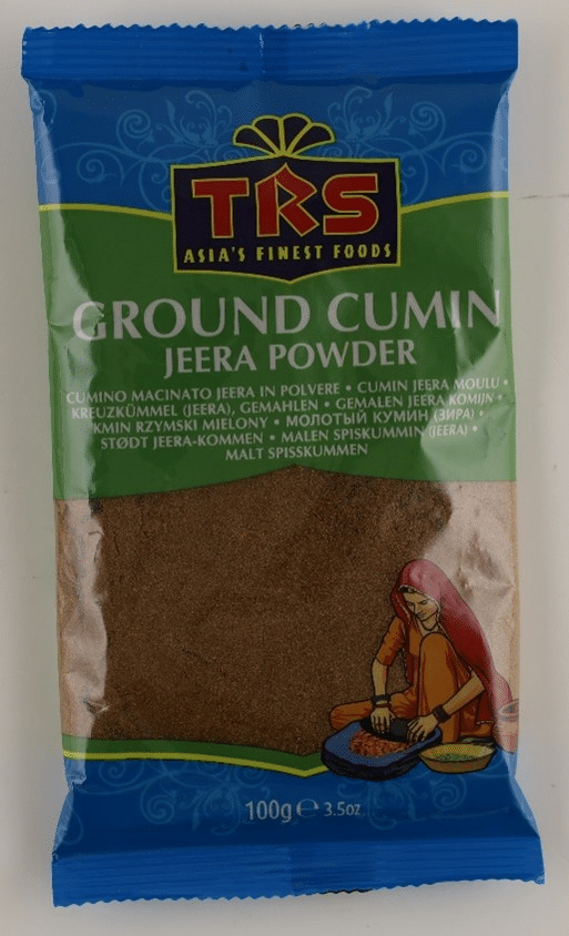 Verpackung des Produkts „TRS Ground Cumin Jeera Powder“. Das Gewürz ist in einer bunt bedruckten Kunststoffverpackung verpackt. Die Vorderseite zeigt den Produktnamen in weißer Schrift auf grünem Hintergrund. Unten rechts ist eine Illustration einer Frau, die Gewürze mahlt. Die Verpackung enthält 100 Gramm gemahlenen Kreuzkümmel.