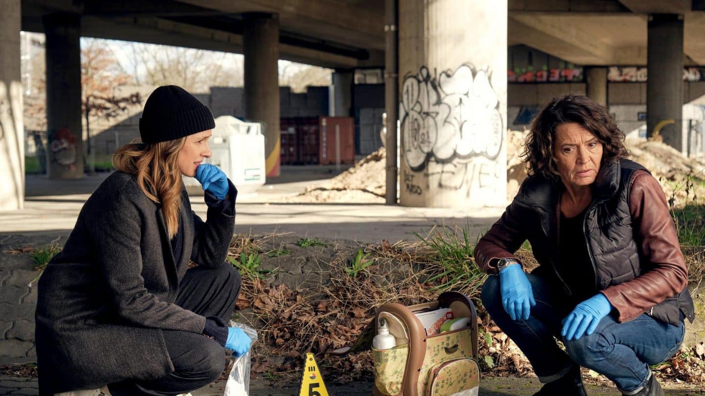 Johanna Stern und Lena Odenthal knien an einem Tatort unter einer Brücke. Beide tragen blaue Handschuhe und wirken nachdenklich. Neben ihnen steht eine Beweismarkierung und eine Tasche mit forensischem Equipment. Im Hintergrund sind Betonpfeiler mit Graffiti sowie Container zu sehen. Die Szene vermittelt einen ernsten und konzentrierten Eindruck.