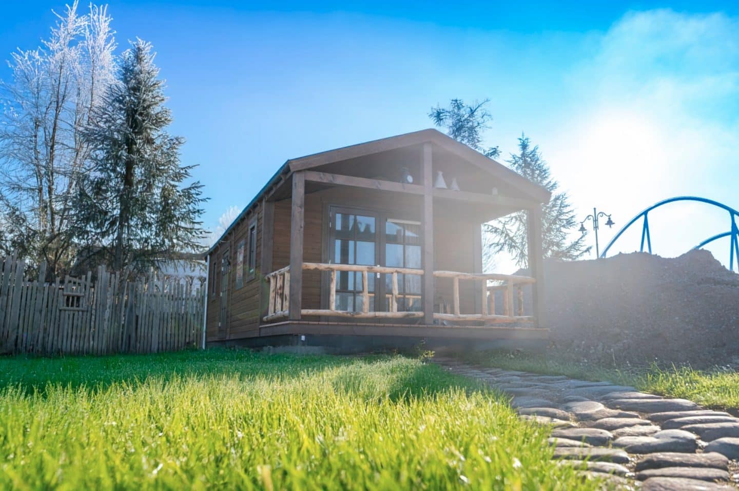Eine rustikale Blockhütte mit überdachter Veranda im Westernstil, umgeben von grüner Wiese und Holzzaun, mit einer Achterbahnstruktur im Hintergrund unter blauem Himmel.
