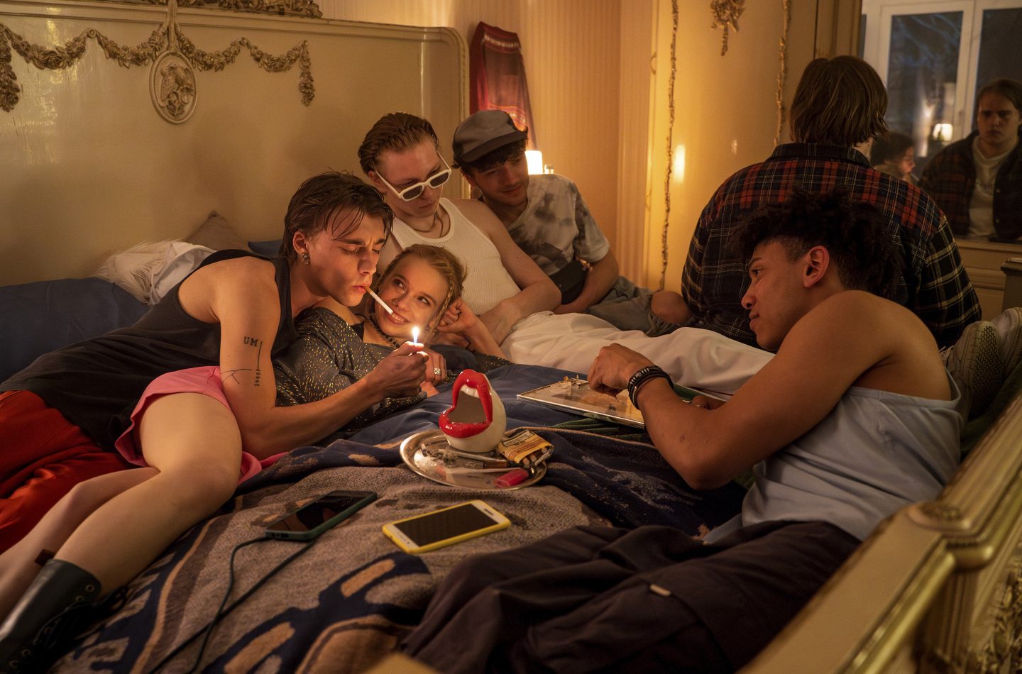Sechs junge Menschen liegen und sitzen entspannt auf einem Bett in einem stimmungsvoll beleuchteten Raum. Einer zündet sich eine Zigarette an, während andere plaudern, lächeln oder ins Spiel vertieft sind. Auf dem Bett liegen Handys, ein Spiegel in Form eines Mundes und andere Partyutensilien.