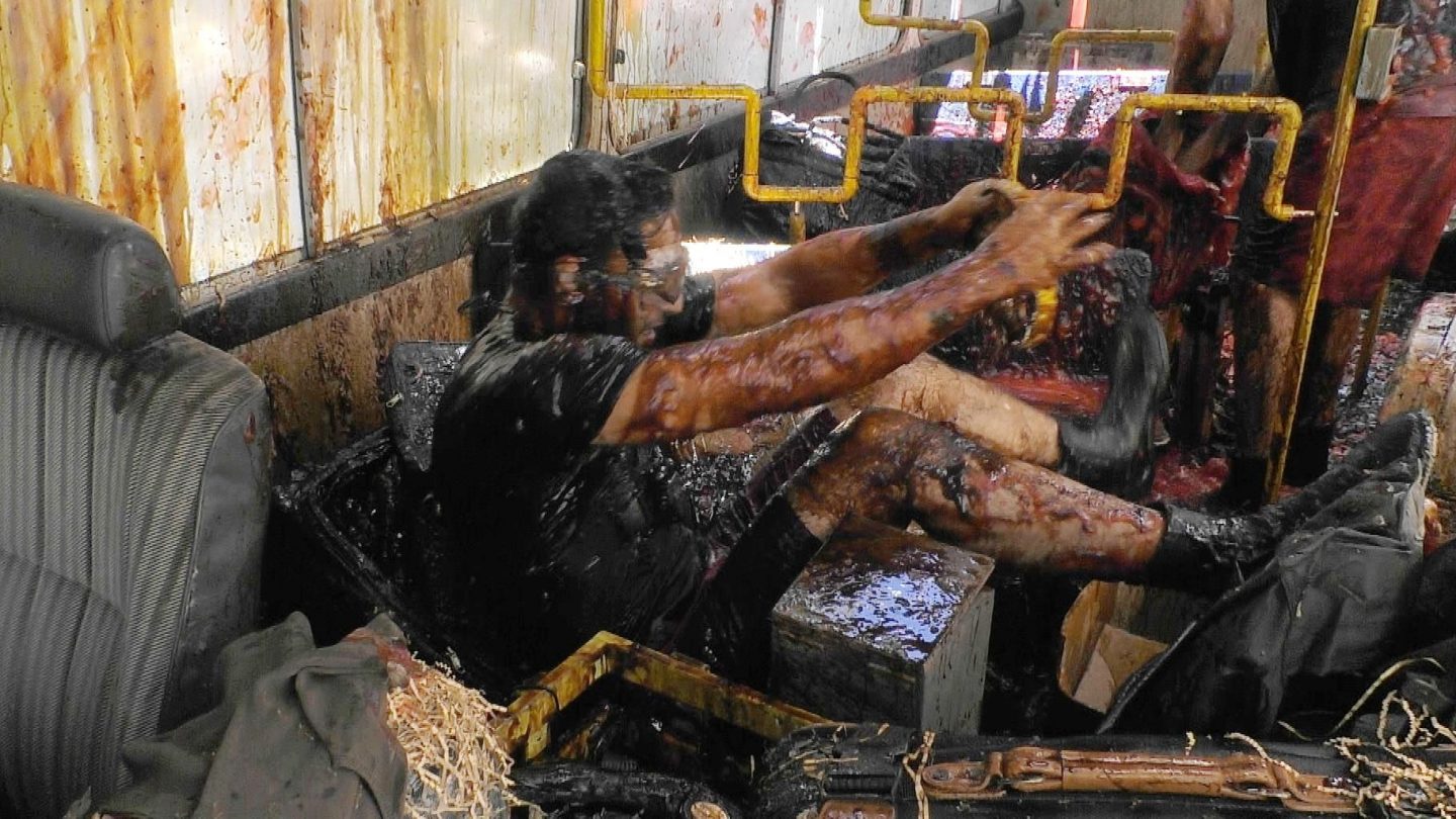 Jörg Dahlmann sitzt in einem völlig verschmutzten Bus, überzogen mit dunklen Flüssigkeiten und Krabbeltieren. Er trägt eine Schutzbrille und hält sich mit den Händen an einer Stange fest, während er sich durch das Chaos kämpft. Der Innenraum ist mit Ekelschleim, Stroh und Insekten bedeckt.