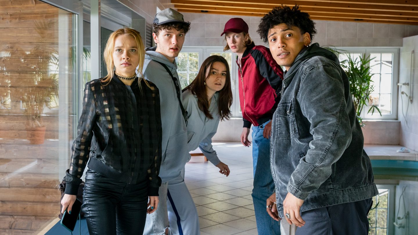 Fünf Jugendliche stehen angespannt in einem modernen Poolhaus mit Glaswänden und Fliesenboden. Maya hält ein Handy in der Hand, während alle mit erschrockenen und überraschten Blicken in eine Richtung schauen. Ihre Körperhaltung wirkt angespannt, als wären sie gerade ertappt worden