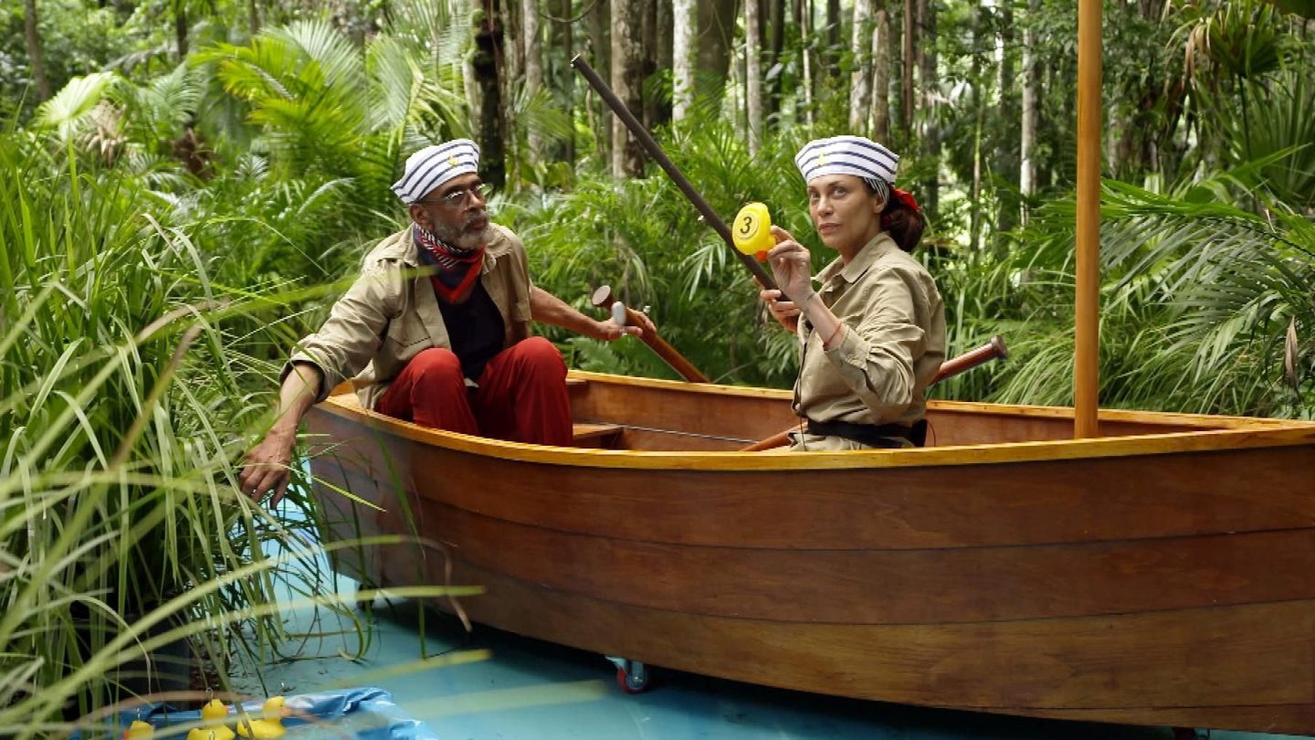 Lilly Becker und Pierre Sanoussi-Bliss sitzen in einem Holzboot mitten im Dschungel und nehmen an der Schatzsuche „Entchen Lotterie“ teil. Lilly hält ein gelbes Plastikentchen mit der Nummer 3 in die Kamera, während Pierre das Boot navigiert. Beide tragen Matrosenmützen und Khaki-Outfits. Im Vordergrund schwimmen weitere Entchen auf einer künstlichen Wasserfläche. Die dichte grüne Vegetation rundherum verstärkt das tropische Abenteuer-Feeling.






