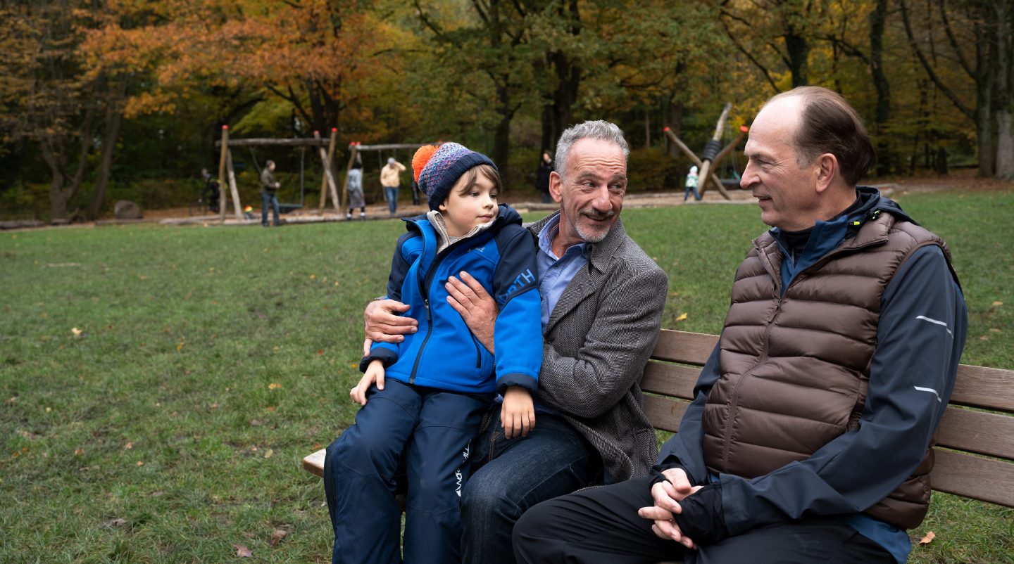 „Markus Gellert (Herbert Knaup, rechts) sitzt mit Jürgen Weber (Heio von Stetten, Mitte) auf einer Parkbank. Jürgen hält seinen Sohn Lion (Heinrich Höhne, links) auf dem Schoß. Der Junge trägt eine blaue Winterjacke und eine Strickmütze. Im Hintergrund ist ein Spielplatz mit mehreren Kindern und Erwachsenen zu sehen. Die Szene spielt im Freien mit herbstlicher Kulisse. Das Bild stammt aus der ARD-Serie 'Die Kanzlei' und zeigt eine Szene aus einem Sorgerechtsstreit.“