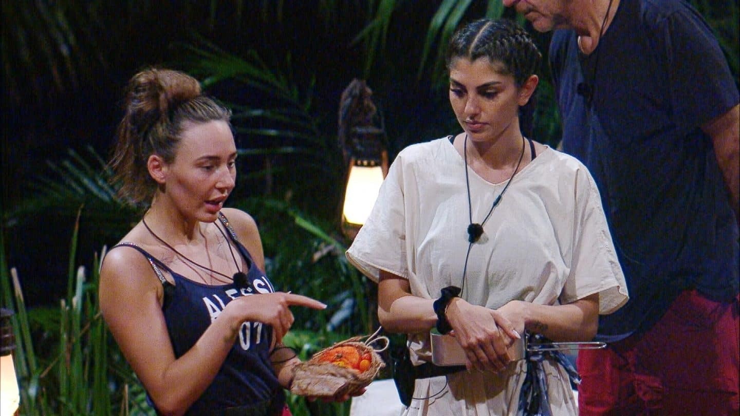 Alessia Herren (links) diskutiert gestikulierend mit Edith (Mitte) über Tomaten, die sie in einer kleinen Tasche hält. Alessia trägt ein schwarzes Top mit Spaghettiträgern, Edith ein weißes lockeres Oberteil mit geflochtenen Haaren. Edith hält ihre Arme verschränkt und schaut ernst. Rechts im Hintergrund steht Yeliz Koc in dunkler Kleidung, während eine Laterne zwischen tropischen Pflanzen leuchtet. Der Streit um die richtige Zubereitung der Tomaten sorgt für Spannung im Camp.