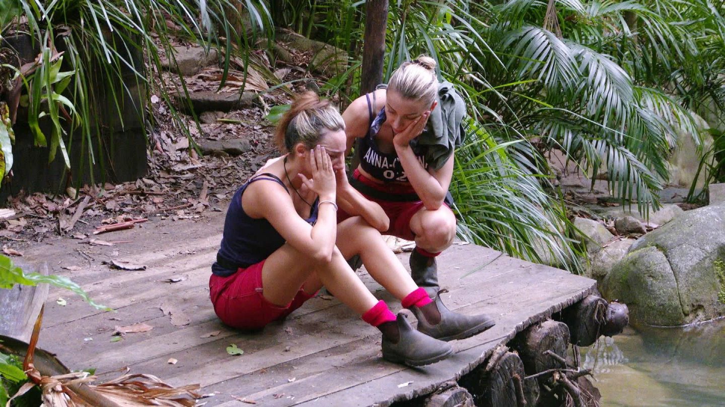 Alessia Herren sitzt mit gesenktem Kopf auf einem Holzsteg am Wasser und hält sich weinend das Gesicht. Neben ihr kniet Anna-Carina Woitschack, die sie mit einer Hand am Gesicht tröstet. Umgeben von tropischer Vegetation wirkt die Szene emotional und nachdenklich.