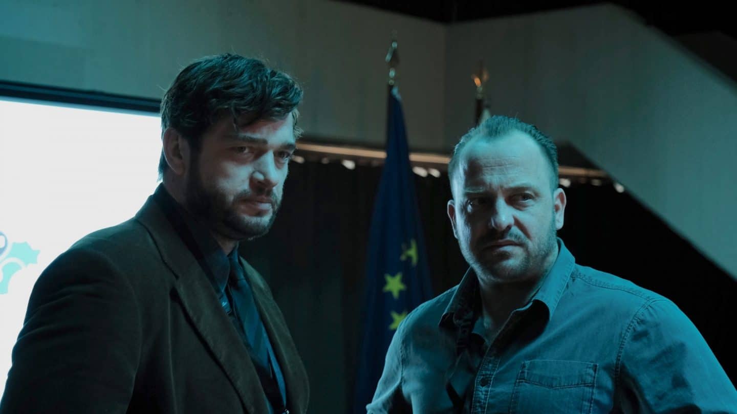 "Das Ende der Wahrheit": Martin Behrens (Ronald Zehrfeld) und Arne Markow (Urs Rechn) stehen nebeneinander und blicken angewidert. Im Hintergrund stehen zwei Fahnenständer. Einer davon trägt die Fahne der Europäischen Union, der andere ist von Arnes Körper verborgen.
