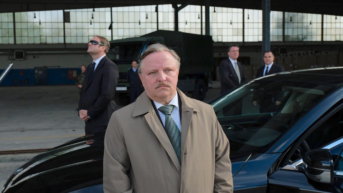 "Das Ende der Wahrheit": Dr. Joachim Rauhweiler (Axel Prahl) in beigem Trenchcoat steht in einer Industriehalle vor einer schwarzen Limousine. Im Hintergrund sieht man mehrere Männer in schwarzen Anzügen.
