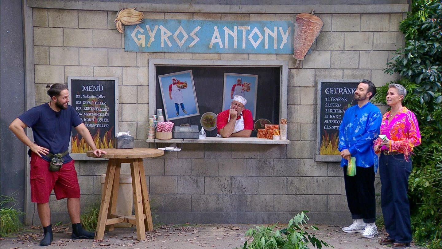 Maurice Dziwak (links) steht entspannt an einem runden Holztisch vor dem Imbiss „Gyros Antony“, bereit für die Dschungelprüfung „Einkaufs-Strafe“. Hinter dem Tresen lehnt ein Mann in rotem Shirt und weißer Schürze nachdenklich auf seiner Hand. Rechts daneben begrüßen die Moderatoren Sonja Zietlow in einer pink-gemusterten Bluse und Jan Köppen in einem blauen Hemd den Kandidaten. Die Kulisse erinnert an eine griechische Imbissbude mit Speisekarten an der Wand.







