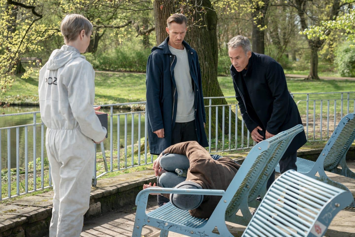 "SOKO Leipzig - Italien kann sehr kalt sein": Prof. Dr. Sabine Rossi (Anna Stieblich), Moritz Brenner (Johannes Hendrik Langer) und Jan Maybach (Marco Girnth) stehen an einer Parkbank, auf der das Opfer Leonard Lübben (Sebastian Stielke) liegt.
