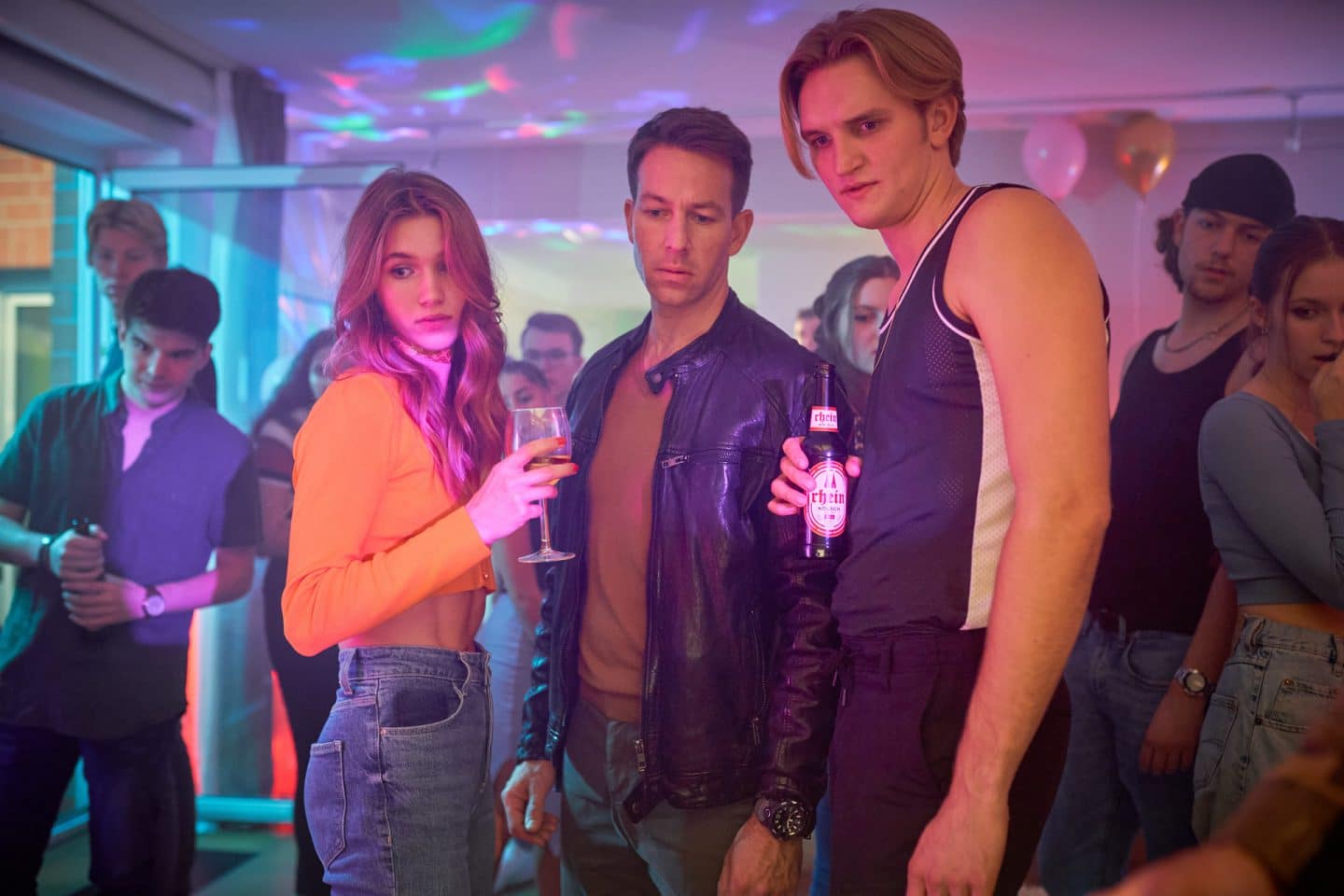 "Marie Brand und die falsche Wahrheit": Isabell Lauterbach (Anna-Lena Schwing), Trainer Victor Holtmann (Florian Anderer) und Jan Frehse (Ludger Bökelmann) stehen zusammen auf der Party und schauen auf einen gemeinsamen Blickpunkt auf dem Boden. Die Partygäste im Hintergrund (Komparsen) schauen ebenfalls dorthin. Isabell hält ein Weinglas in der Hand und Jan eine Bierflasche.