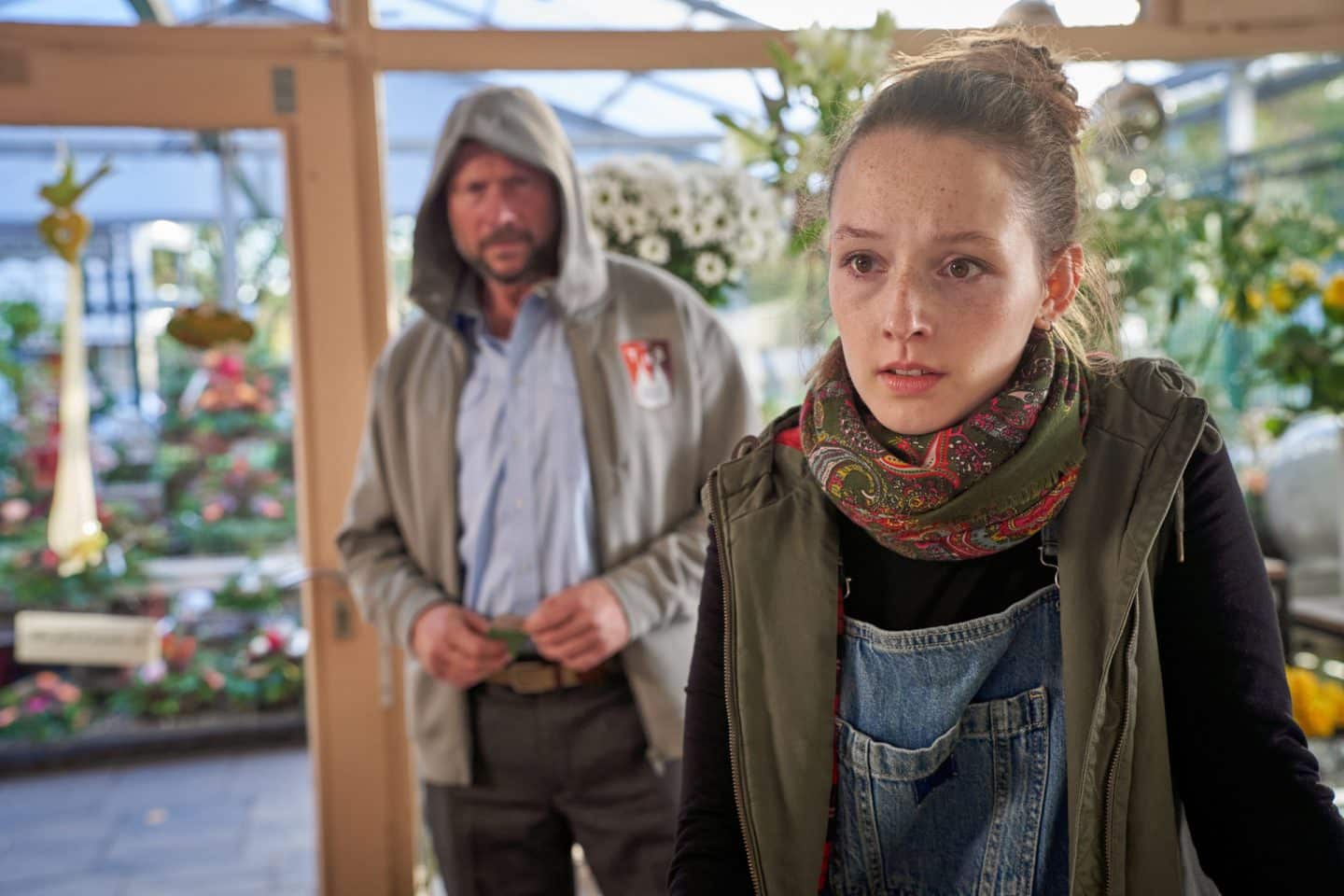 "Marie Brand und die falsche Wahrheit": Melanie Josten (Nora Solcher) steht geschockt in ihrer Gärtnerei am Tresen und sieht zum linken Bildrand. Im Hintergrund steht Klaus Thiele (Hanno Friedrich) mit einem grauen Kapuzenpullover und blickt zu Melanie Josten.