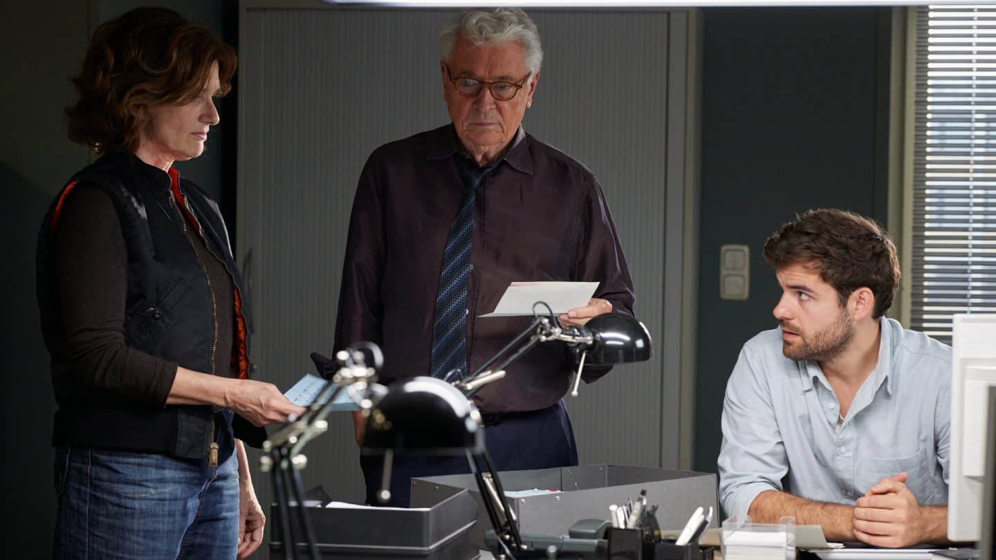 "Die Stille am Ende der Nacht": Johannes Fischer (Henry Hübchen) steht zwischen Konstanze Satorius (Victoria Trauttmansdorff) und Jan Kettler (Lucas Reiber) in einem Büro und blickt fragend nach unten, während er einen Brief in der Hand hält. Konstanze Satorius hält ebenfalls einen Brief in der Hand und schaut zu Jan Kettler, der zurück zu ihr schaut.
