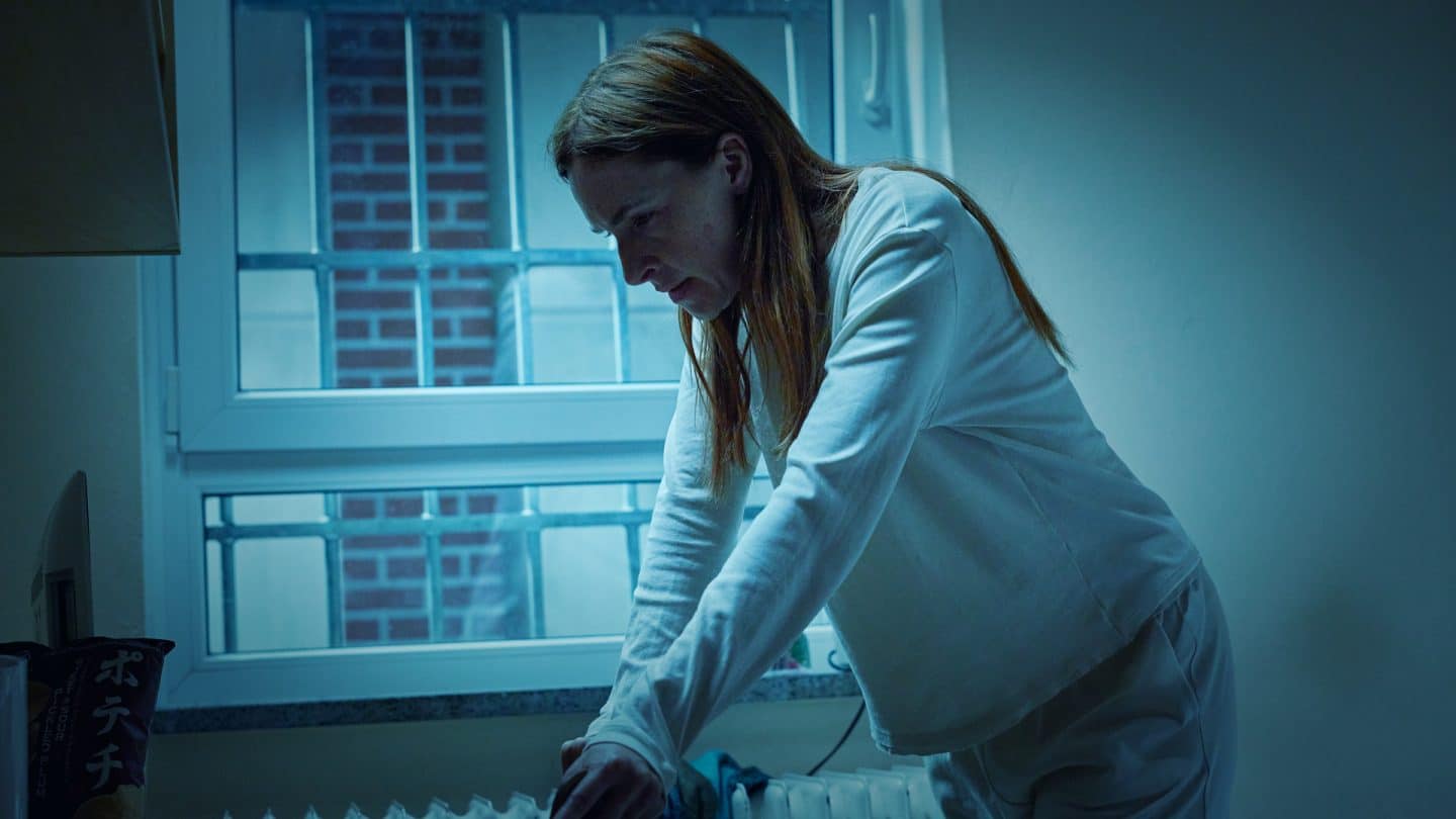 "Die Stille am Ende der Nacht": Liane Sievers (Kim Riedle) steht in einem dunklen Raum mit Gittern hinter dem Fenster und trägt Häftlingskleidung. Sie blickt verzweifelt nach unten und hält sich an einem Stuhl fest.
