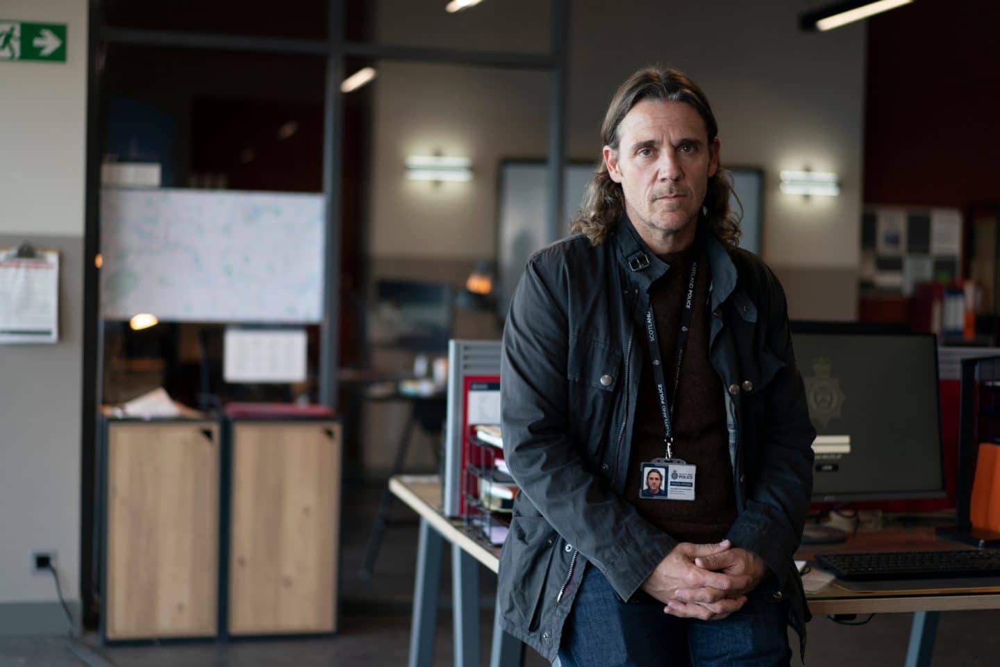 "Annika – Mord an Schottlands Küste - Der Seehund": DS Michael McAndrews (Jamie Sives) sitzt im Büro auf der Kante eines kleinen Schreibtisches, auf dem Computer stehen.
