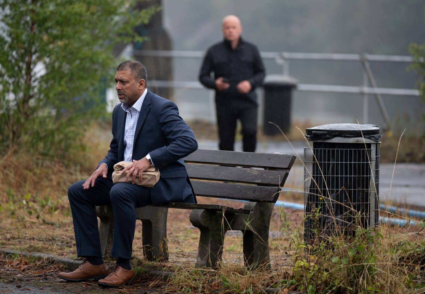 "Annika – Mord an Schottlands Küste - Der Seehund": Rihan Harijan (Pal Aron) sitzt im Freien auf einer Bank. Er hält eine Papiertüte mit der linken Hand fest und wirkt angespannt. Im Hintergrund taucht Jaden Stanton (Martin Pemberton) auf.
