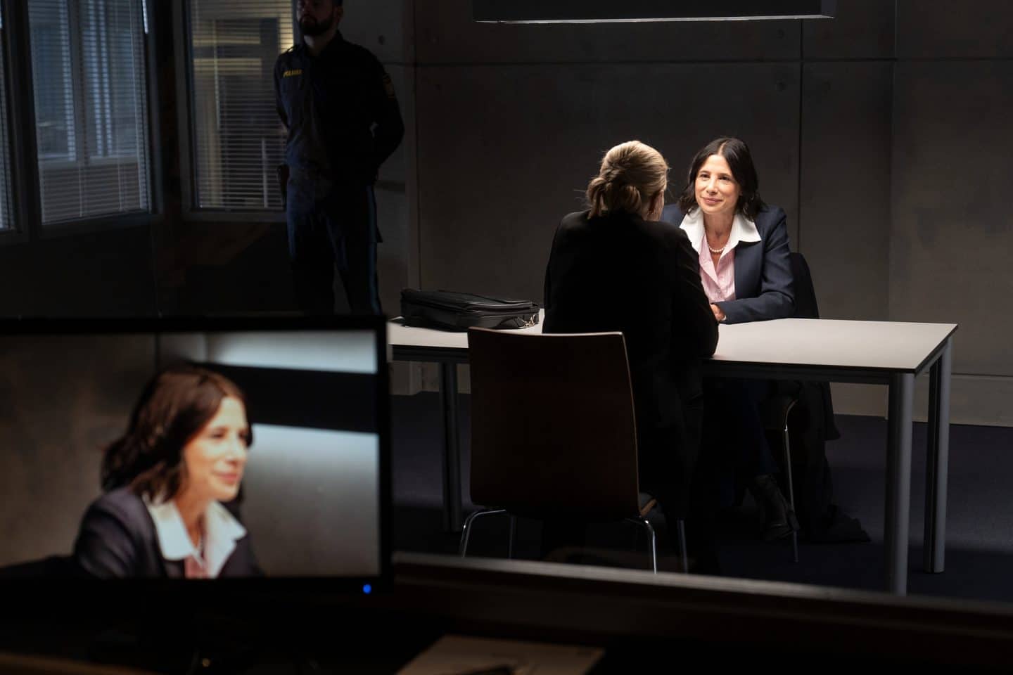 "Die Chefin - Fake News": Eine Glasscheibe gibt den Blick ins abgedunkelte Verhörzimmer frei, in welchem Demet Palak (Isabella Parkinson) hinter dem Tisch sitzt und überlegen ihr Gegenüber anlächelt, welches mit dem Rücken zur Kamera sitzt. Vor der Glasscheibe steht ein Monitor, der Palak in Nahaufnahme zeigt.
