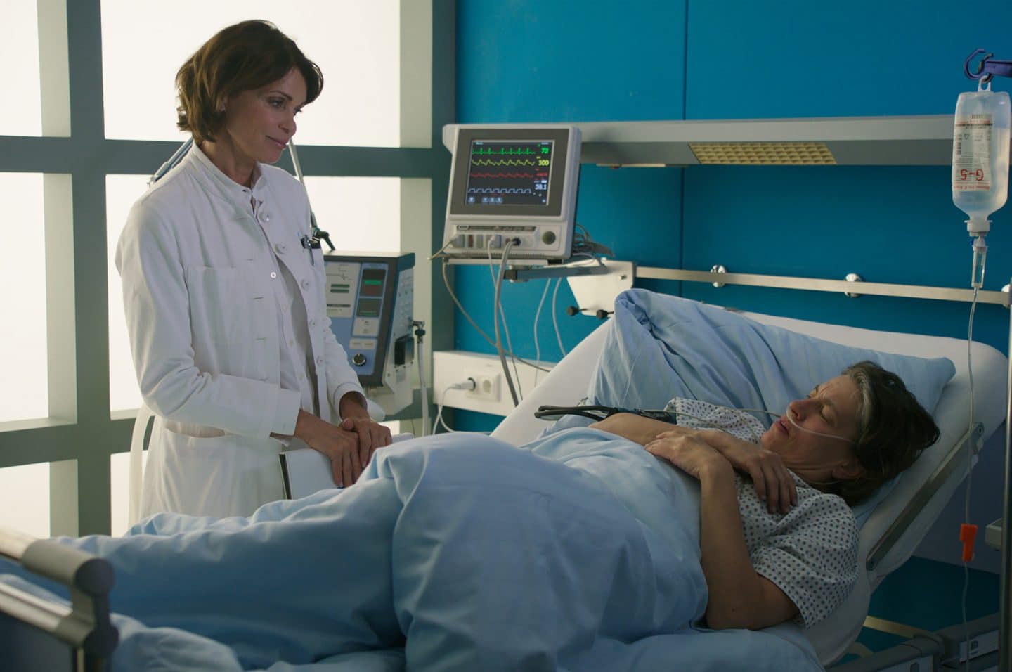 "Notruf Hafenkante - Ausgebrannt": Dr. Jasmin Jonas (Gerit Kling) steht neben dem Bett von Rieke Clemens (Jule Gartzke) im Krankenzimmer des EKH. Sie grinst sie freundlich an.
