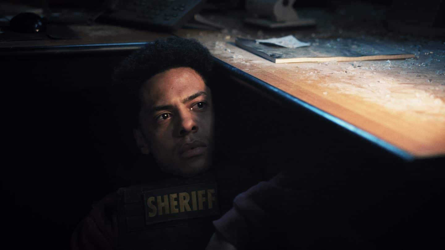 "Jericho Ridge - Unter Beschuss": Monty (Zack Morris) kauert unter einem Tisch, auf dem Glasscherben verteilt sind. Er schaut darunter hervor und sieht verängstigt aus.
