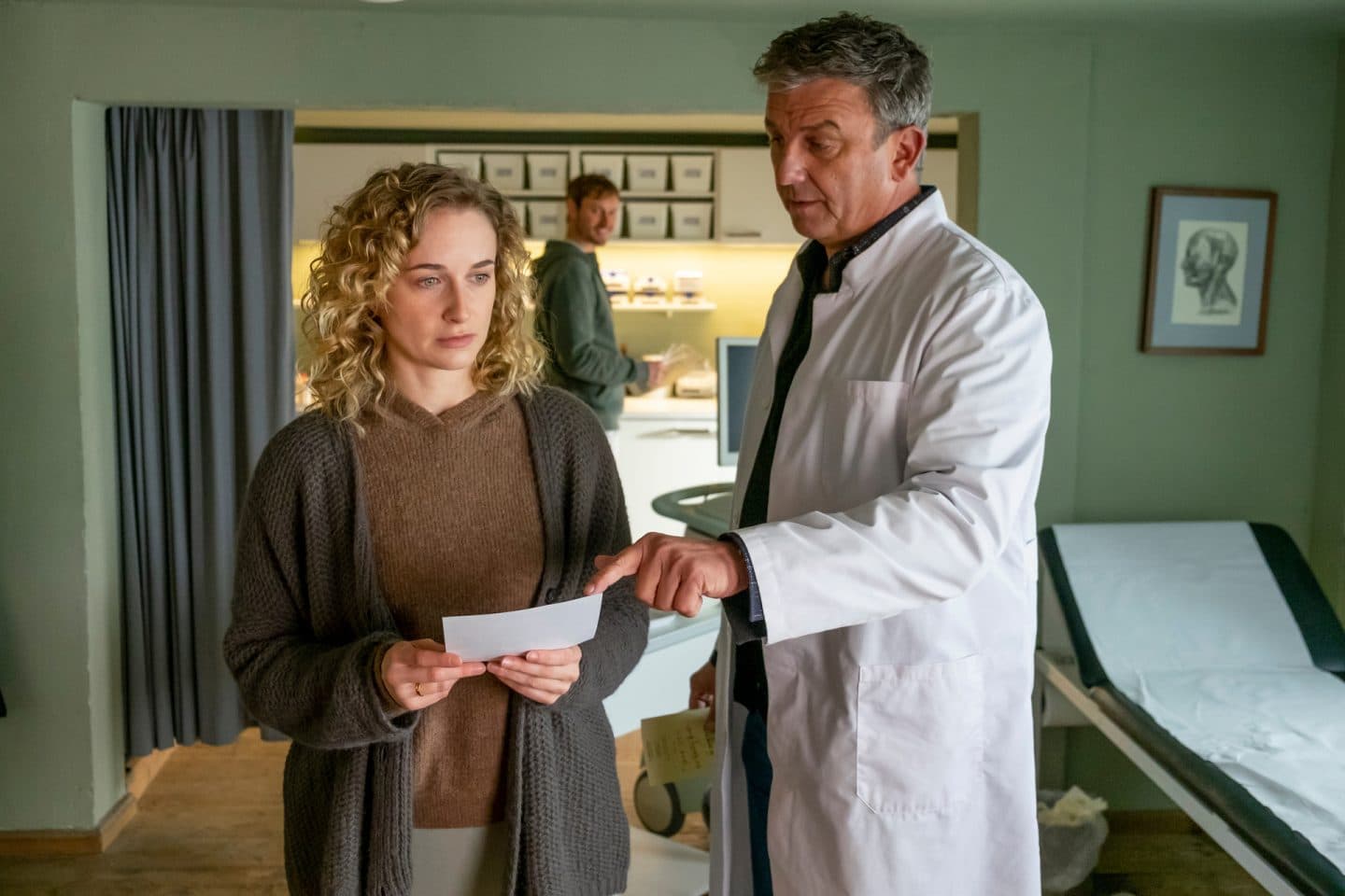 "Der Bergdoktor: Sternenkinder": In der Arztpraxis: Nachdenklich hält Jana Weiss (Pia Barucki) ein Foto in den Händen. Dr. Martin Gruber (Hans Sigl), der neben ihr steht, zeigt mit dem Finger darauf. Im Hintergrund steht David Kästner (Frédéric Brossier) und beobachtet die beiden.
