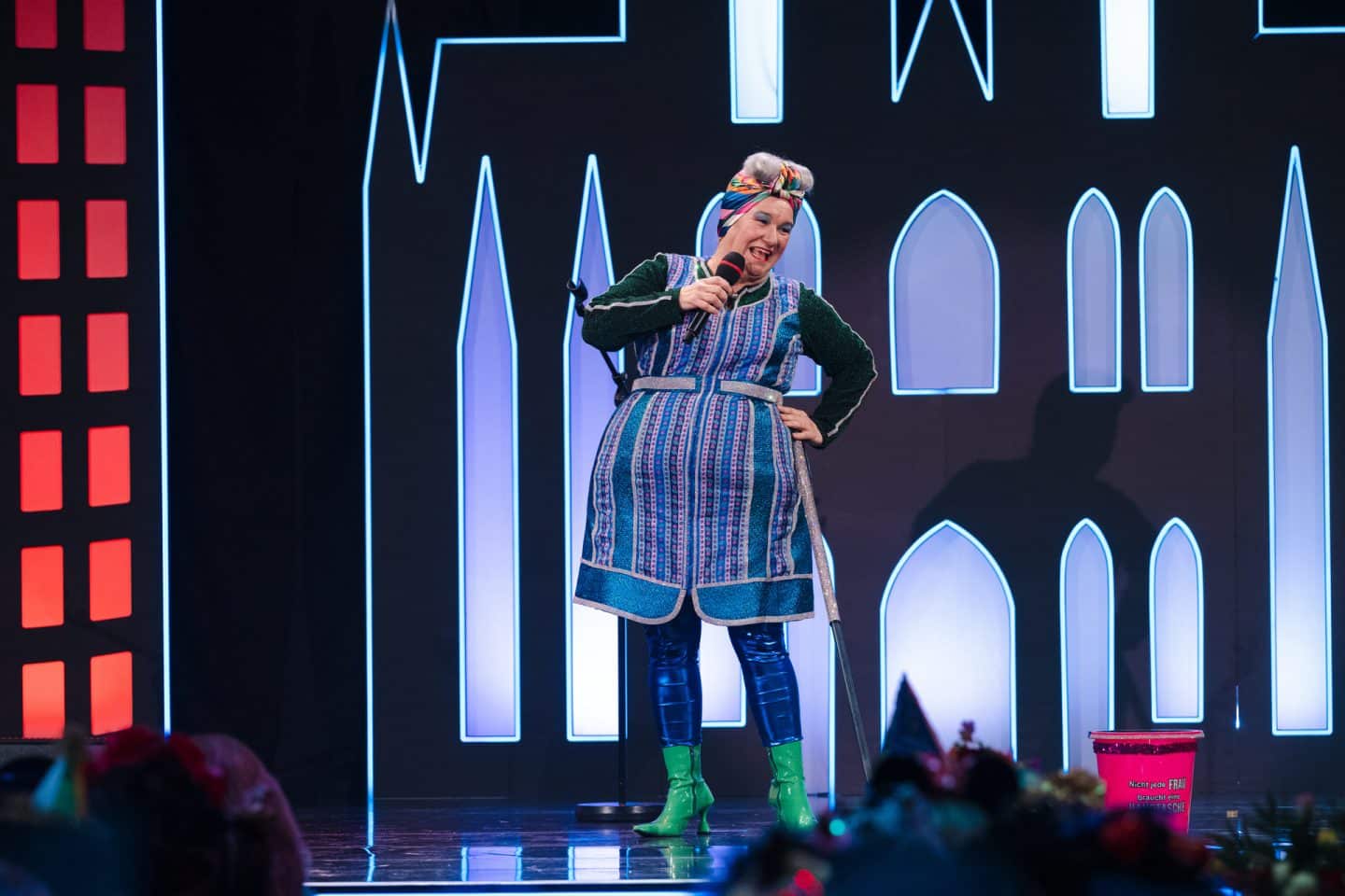 "Kölle Alaaf - Die Mädchensitzung": Rednerin Achnes Kasulke (Annette Esser) steht als Putzfrau auf der Bühne. In der Hand hält sie ein Mikrofon, sie beugt sich leicht zur Seite und lächelt in die Kamera.
