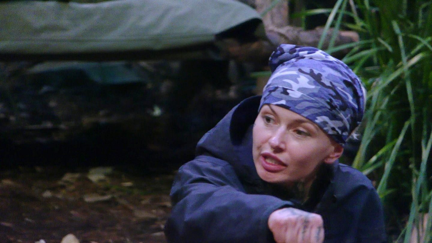 Edith Stehfest sitzt mit entschlossenem Blick im Dschungelcamp und gestikuliert energisch während einer hitzigen Diskussion. Sie trägt eine dunkle Jacke und ein blau-graues Camouflage-Bandana, ihre tätowierte Hand ist deutlich zu sehen. Der Hintergrund ist düster, das Camp-Setting mit Pflanzen und Holzstrukturen verstärkt die angespannte Atmosphäre. Ihre Mimik zeigt Entschlossenheit und Emotion – sie verteidigt ihre Position in der Auseinandersetzung.






