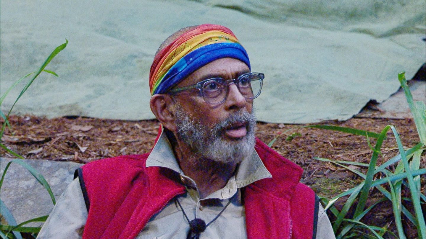 Pierre Sanoussi-Bliss sitzt im Dschungelcamp mit überraschter und ungläubiger Miene. Er trägt eine Brille, eine rote Weste über einem beigen Hemd und ein buntes Stirnband in Regenbogenfarben. Die Umgebung ist von Pflanzen und Waldboden geprägt. Seine Reaktion deutet auf eine hitzige Diskussion hin, während er ruhig, aber bestimmt auf eine Situation im Camp reagiert.






