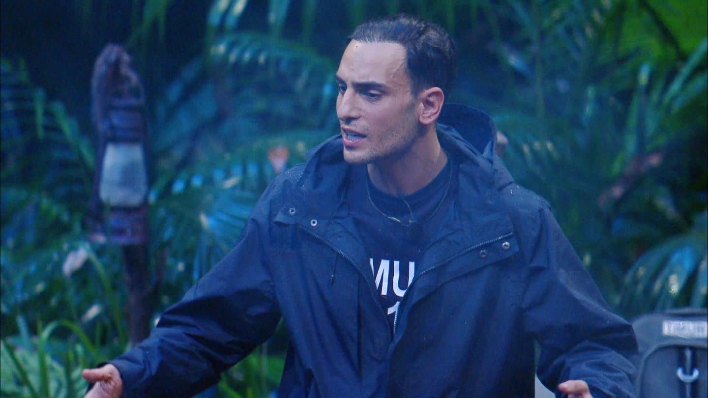 Timur Ülker steht mit ernster Miene und ausgebreiteten Armen im Dschungelcamp, während er in einer hitzigen Diskussion spricht. Er trägt eine dunkle Regenjacke, sein Gesichtsausdruck zeigt Entschlossenheit und Emotion. Der grüne Dschungelhintergrund mit Laterne und Pflanzen verstärkt die dramatische Atmosphäre des Moments, in dem er am Lagerfeuer eine Debatte entfacht.






