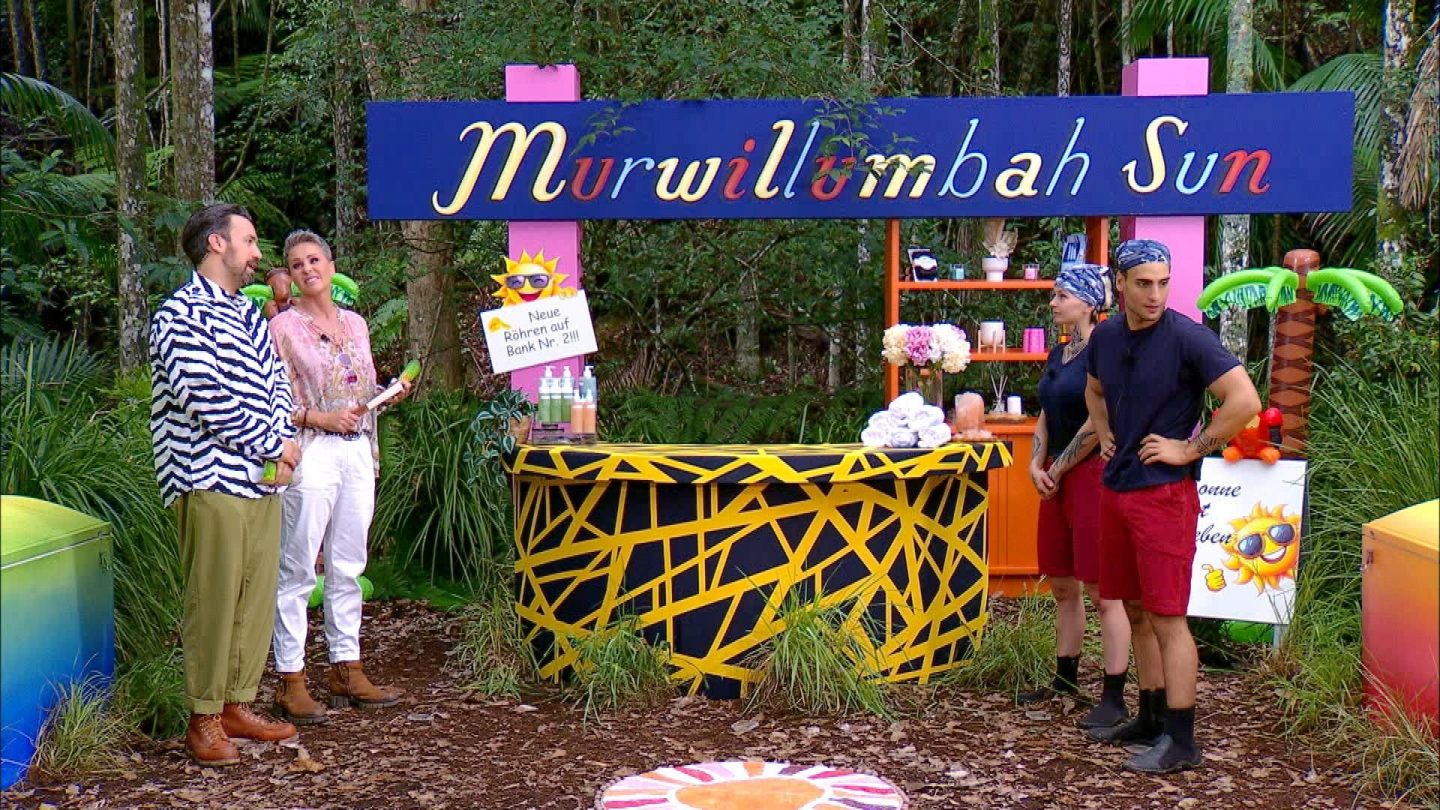 Timur Ülker und Edith Stehfest stehen angespannt vor der Dschungelprüfung „Murwillumbah Sun“, bereit für ihre Herausforderung. Die farbenfrohe Kulisse mit Sonnenstudio-Dekoration und künstlichen Palmen wirkt täuschend idyllisch. Links im Bild beobachten die Moderatoren Sonja Zietlow und Jan Köppen das Geschehen, Sonja mit einem Lächeln, Jan im auffälligen Zebra-Hemd. Auf dem Tresen liegen Handtücher und Flaschen, während im Hintergrund ein Schild auf neue Röhren hinweist. Die Prüfung verspricht Nervenkitzel – doch werden Timur und Edith diesmal Sterne fürs Camp holen?