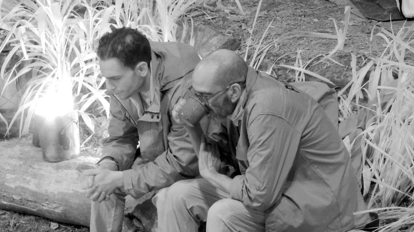 Timur Ülker (links) und Pierre Sanoussi-Bliss sitzen in der Nachtwache des Dschungelcamps vor dem Lagerfeuer. Timur wirkt nachdenklich, die Hände gefaltet, während Pierre aus einem Blechbecher trinkt. Die Szene ist in Schwarz-Weiß gehalten, wodurch die angespannte und reflektierte Stimmung verstärkt wird. Eine Laterne wirft Licht auf das Geschehen, während das Camp im Hintergrund in Dunkelheit getaucht ist.