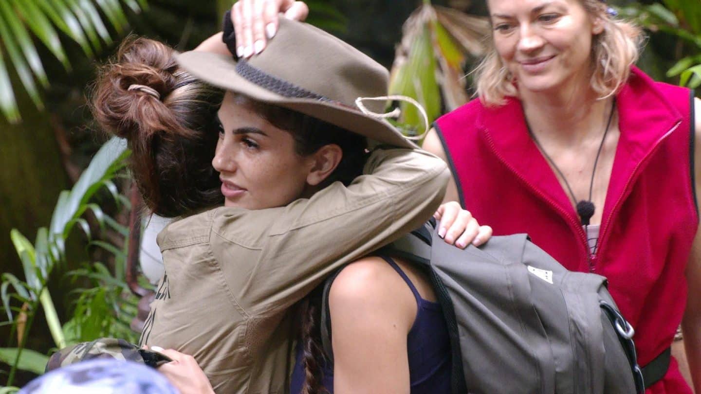 Yeliz Koc mit olivgrünem Hut und Rucksack verabschiedet sich im Dschungelcamp. Sie umarmt eine Mitbewohnerin, während eine weitere Camperin in roter Weste lächelnd danebensteht. Die Zuschauer haben entschieden: Yeliz muss das Camp verlassen.







