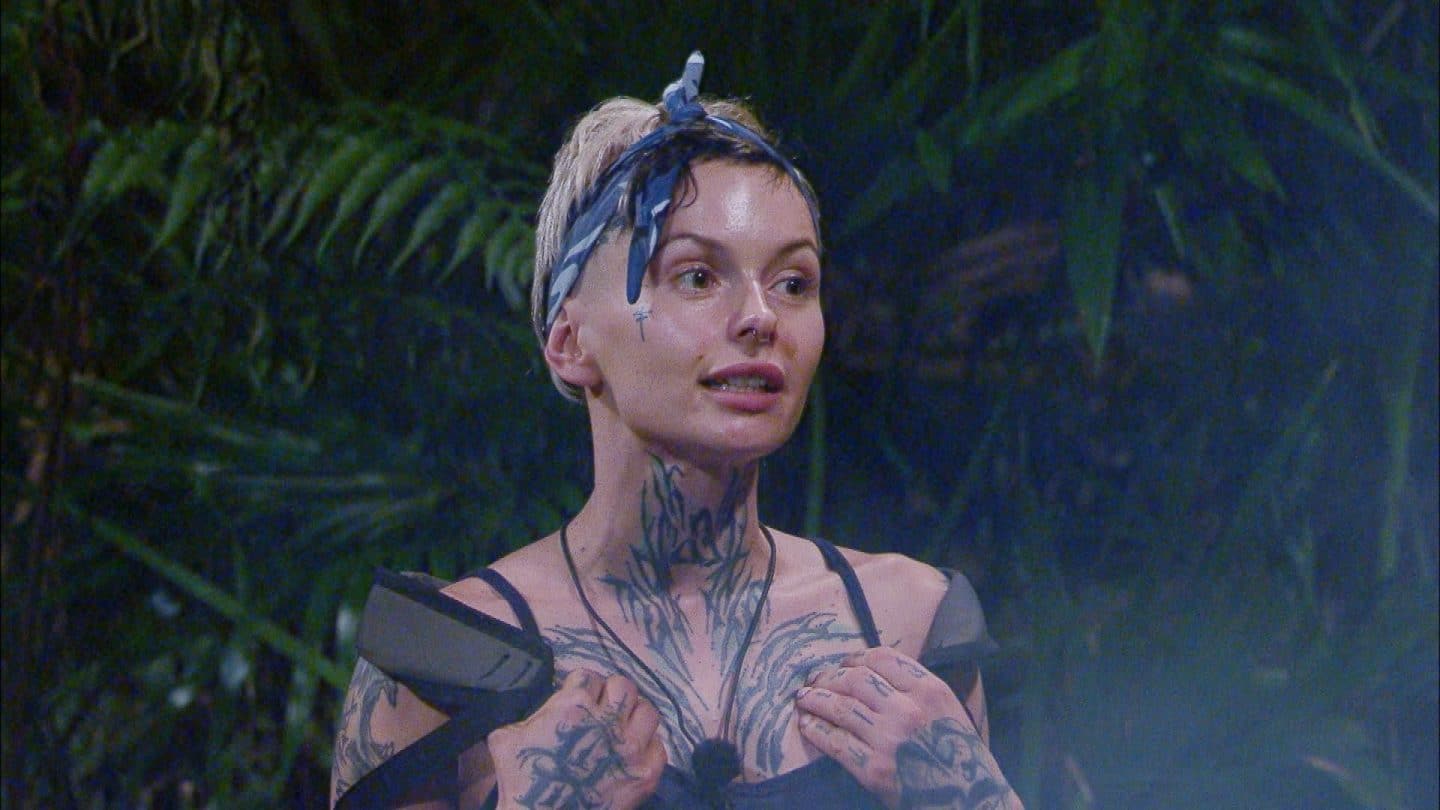 Edith Stehfest steht mit entschlossener Miene im Dschungelcamp, umgeben von tropischer Vegetation. Die tätowierte Musikerin trägt einen blauen Kopfschmuck und wirkt angespannt, während sie ihre Hände an ihre Schultern legt.