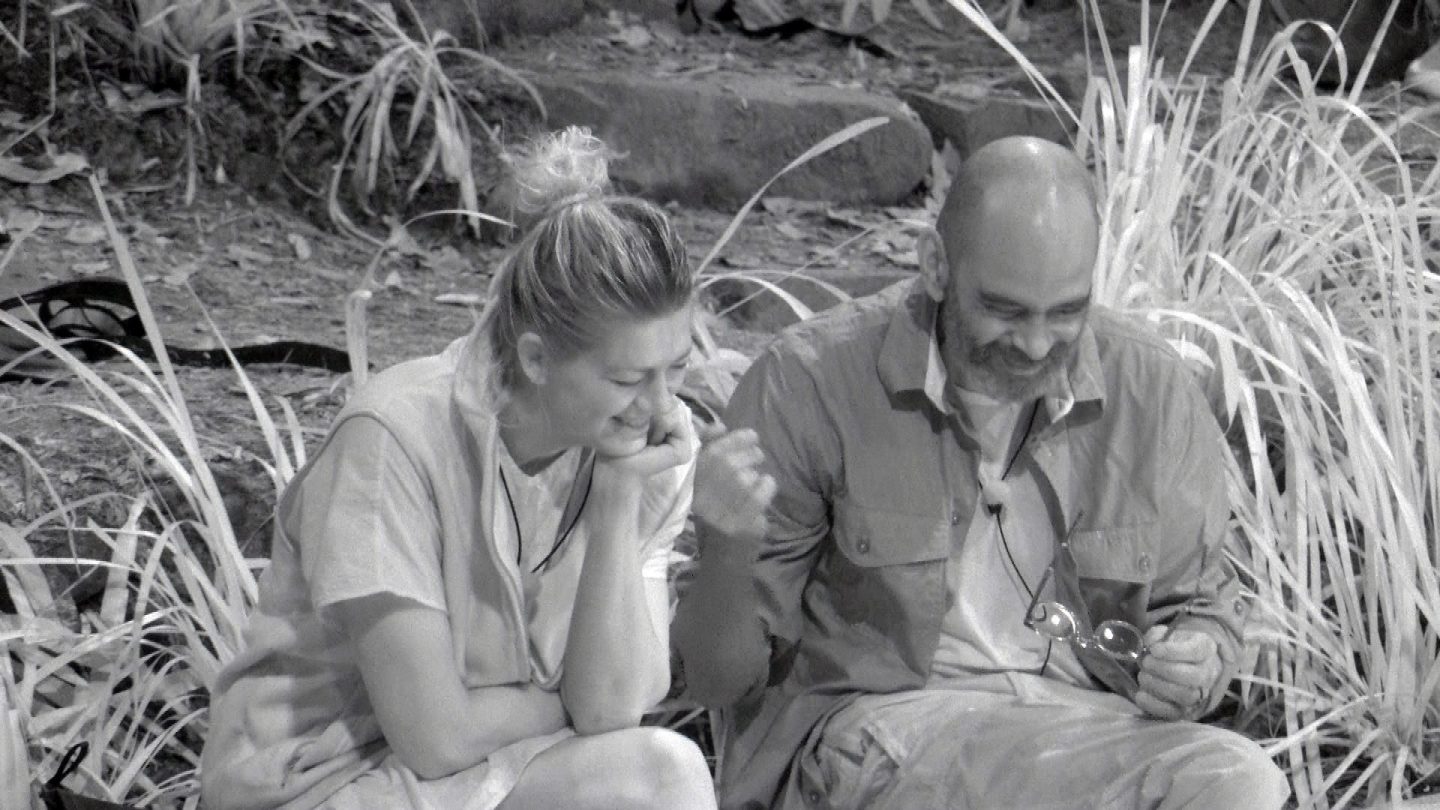 Schwarz-Weiß-Aufnahme von Nina Bott und Pierre Sanoussi-Bliss im Dschungelcamp. Beide sitzen nebeneinander, lächeln und wirken in ein Gespräch vertieft. Pierre hält eine Brille in der Hand, während Nina ihren Kopf auf ihre Hand stützt.