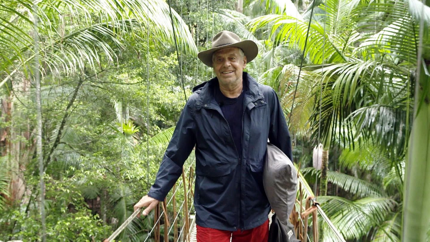 Jürgen Hingsen läuft über eine Hängebrücke im Dschungel, umgeben von grünen Palmen. Er trägt eine dunkle Jacke, einen Hut und hält seinen Schlafsack in der Hand. Mit einem Lächeln verabschiedet er sich vom Camp.