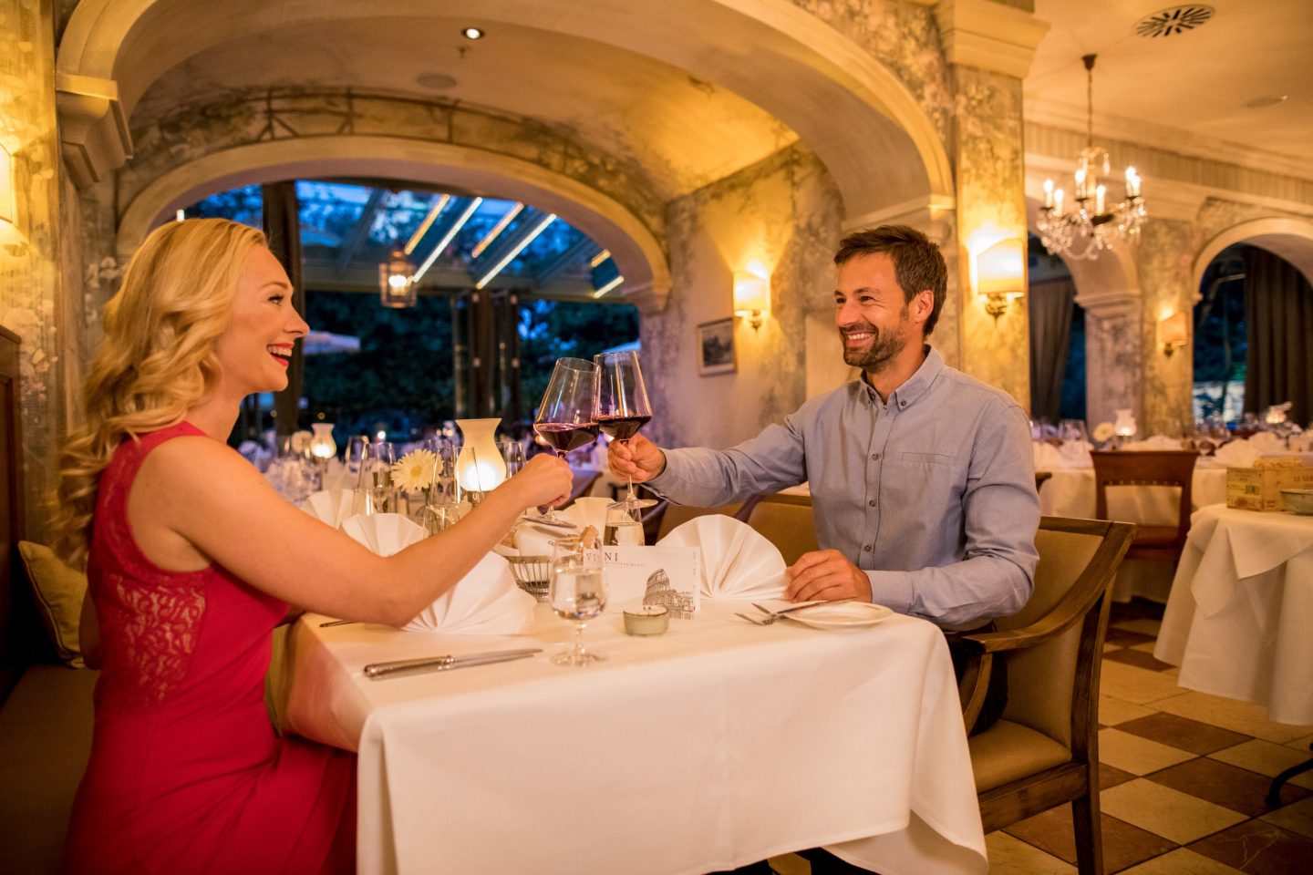 Ein verliebtes Paar stößt mit Rotweingläsern in einem romantisch beleuchteten Restaurant an. Die Frau trägt ein elegantes rotes Kleid, der Mann ein hellblaues Hemd. Die stilvolle Atmosphäre wird durch Kerzenlicht, festlich gedeckte Tische und eine luxuriöse Inneneinrichtung mit Kronleuchtern unterstrichen.