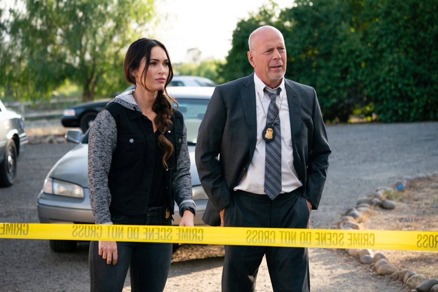 "Die Spur des Killers – Midnight in the Switchgrass": FBI-Agent Karl Helter (Bruce Willis) in Anzug, Krawatte und mit einem FBI-Abzeichen um den Hals, sowie seine Kollegin Rebecca Lombardi (Megan Fox) in Zivil stehen im Freien vor einem von der Polizei abgesperrten Bereich. Im Hintergrund stehen diverse Autos.