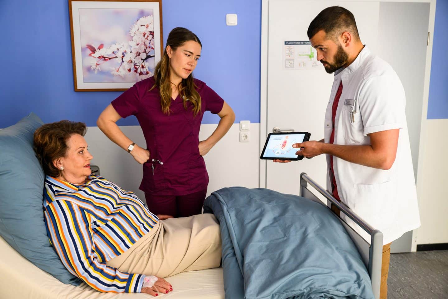 "Bettys Diagnose - Verfahren": Dr. Tarik Şahin (Nicolas Fethi Türksever) zeigt der im Krankenbett liegenden Isabella Schulte (Swetlana Schönfeld) auf dem Tablet seine Diagnose. Betty (Henrike Hahn) steht am Bett der Patientin, sie hat die Hände in die Hüften gestemmt. Im Hintergrund hängt ein Blumenbild an der Wand.
